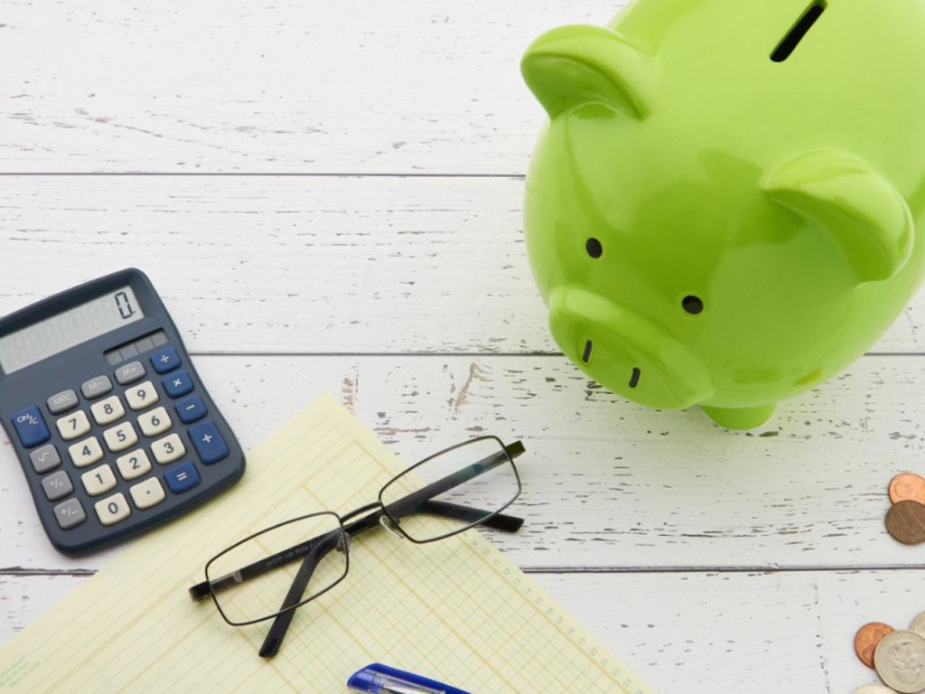Green piggy bank on a white wooden table beside a calculator, notepad, glasses and a few coins