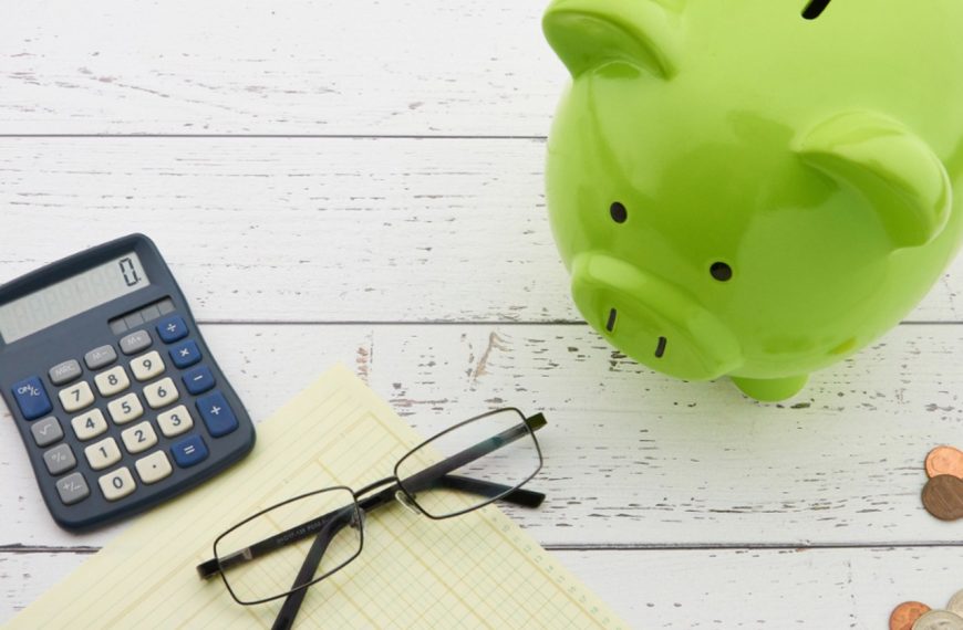 Green piggy bank on a white wooden table beside a calculator, notepad, glasses and a few coins