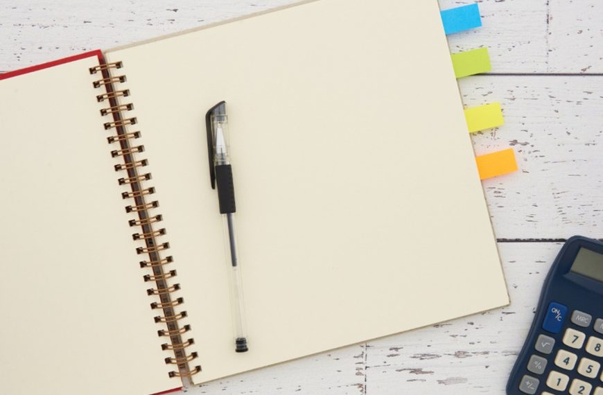 Open spiral notebook with a pen, colored page tabs and a calculator on a white wooden table