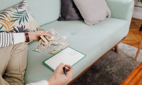 Adult counting cash and writing in a notebook while seated on a stylish couch at home.