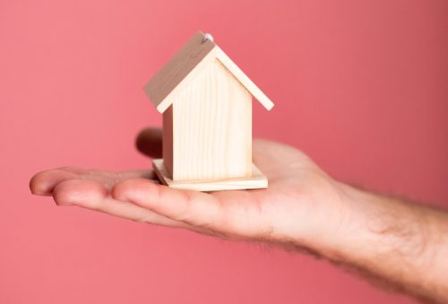 Wooden house model in hand symbolizing real estate concepts on pink background.